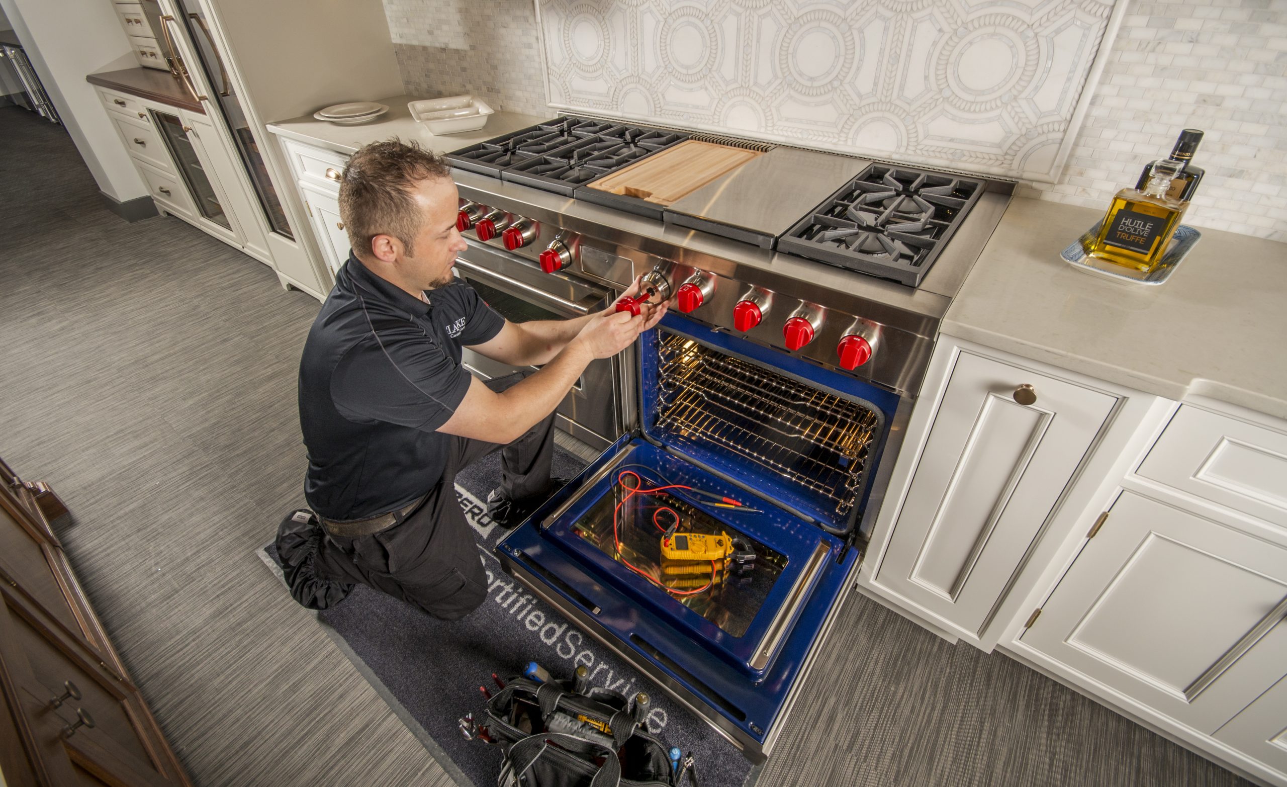 A technician kneels in front of an open oven, using a tool to adjust the knobs while repair equipment is placed on the floor and inside the oven—demonstrating BEST appliance service expertise.