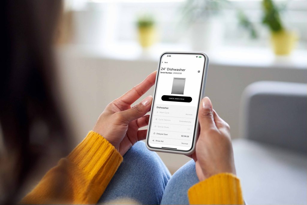 Person holding a smartphone displaying an app interface for managing a 24" dishwasher, featuring options to control and monitor the appliance—ideal for seamless wolf appliance service in any luxury kitchen.