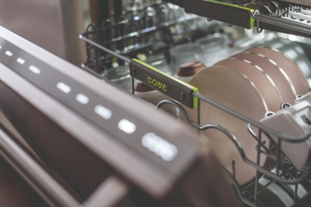 Open dishwasher filled with dishes and cookware; the brand name "Cove" is visible on the green-accented racks.