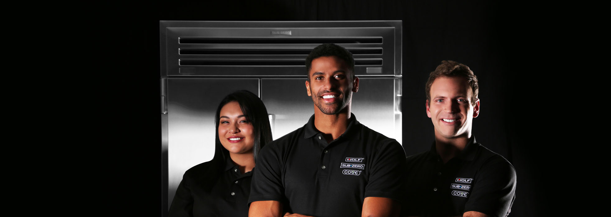 Three people in matching black polo shirts stand in front of a large stainless steel appliance, smiling, as if ready to demonstrate how to clean the filter on a Cove dishwasher, against a sleek black background.