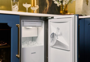 An under-counter ice maker with the door open reveals a bin filled with ice cubes; a clear scoop hangs on the inside of the door.